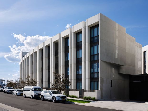 Street-level exterior view of a contemporary multi-storey urban building with a strongly repetitive facade. Tall recessed window bays are framed by projecting pale concrete vertical fins, with dark metal balcony railings and large glazed openings. A solid projecting concrete volume and dark entry/garage element anchor the right side, while parked vehicles and a paved footpath line the street.