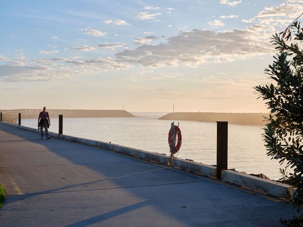 A waterfront concrete promenade or pier viewed at sunset, with calm water extending toward a narrow opening between sandy breakwaters. A person walks along the edge, and a life ring is mounted on the low barrier. Vegetation frames the right side, while the warm sky and distant shoreline create an open coastal atmosphere.