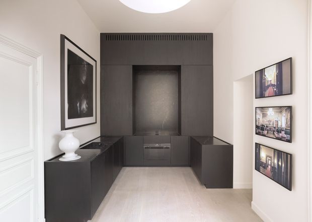 A compact, refined interior hallway or entry zone with dark built-in cabinetry arranged around a recessed central niche, likely incorporating a concealed fireplace or display area. The space has pale timber flooring, white paneled walls and a white door, a large framed monochrome artwork, three framed images, and a circular ceiling light. The cabinetry has a sleek flat-panel appearance with integrated ventilation detailing and a dark stone-like benchtop.