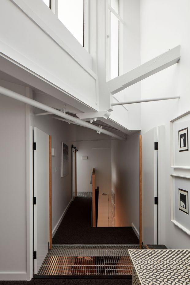 A narrow, bright interior landing or hallway viewed from an elevated level, with white painted walls, exposed white structural beams and pipes, tall glazing, black carpeted flooring, and a metal-grate floor section over a lower level. Open white doors with timber edges lead along the corridor, while framed monochrome artwork and a patterned surface appear at the right. The space has a utilitarian yet carefully renovated character with strong vertical circulation cues.