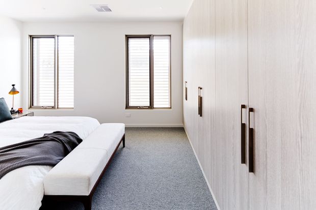 Bright, contemporary bedroom with a large upholstered bed, dark throw, grey carpet, white walls, two timber-framed windows with blinds, and a full-height wall of pale timber built-in wardrobes with dark vertical handles. The room has a clean, restrained, light-filled appearance.