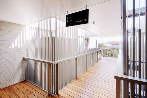 A bright, modern multi-level circulation space with a timber-lined floor, white-painted brick wall, and extensive vertical white metal screening forming balustrades and partitions. A suspended black directional sign hangs above the landing, while large windows and open glazing bring in abundant daylight and provide views toward neighbouring buildings.
