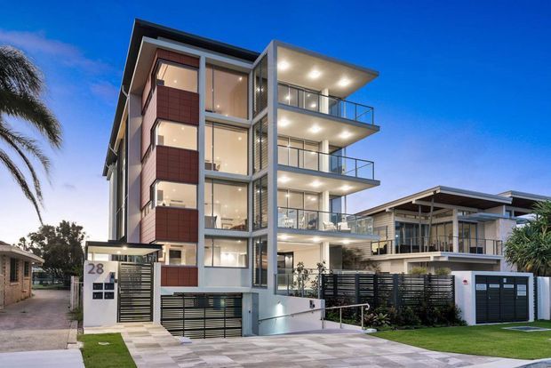 Twilight exterior view of a contemporary multi-storey residential apartment building with prominent floor-to-ceiling glazing, projecting balconies, dark metal balustrades, grey rendered walls, and vertical reddish-brown feature panels. A driveway, garage access, landscaped frontage, and an adjacent modern low-rise residence are visible.