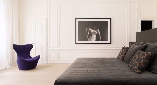 Elegant bedroom with a low-profile dark upholstered bed in the foreground, layered charcoal and patterned cushions, pale timber flooring, and white walls featuring ornate raised panel moulding. A sculptural purple lounge chair sits beside sheer curtains, while a monochrome framed artwork adds a refined focal point.