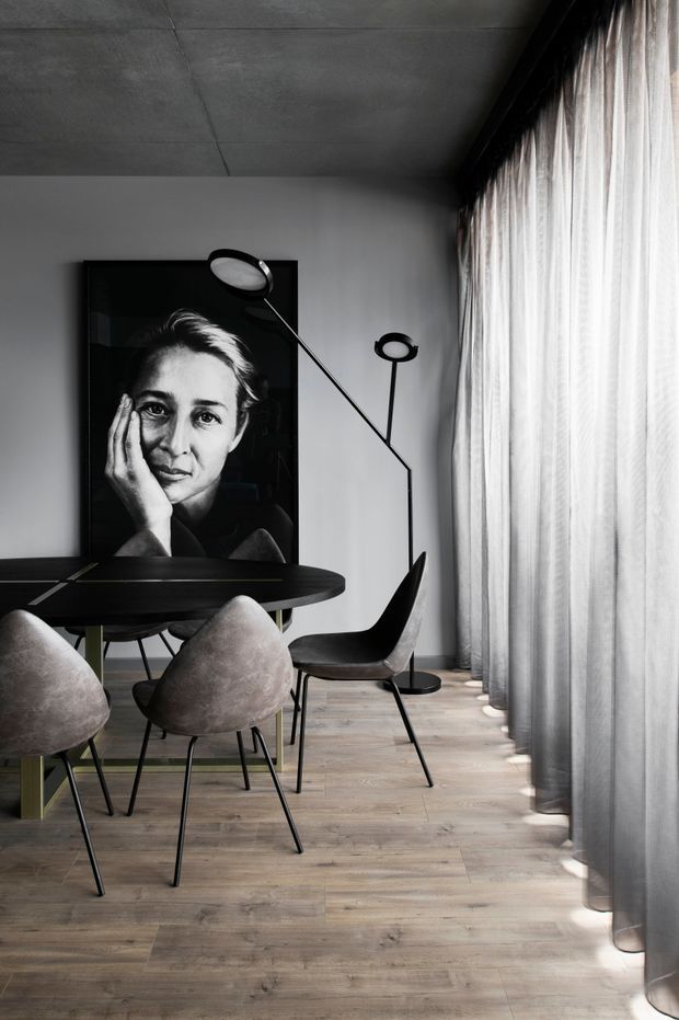 A refined monochromatic dining area with a round black dining table, sculptural upholstered dining chairs, and slender black floor lamps. A large black-and-white portrait leans against the white wall, while full-height sheer curtains line the right side, creating soft filtered daylight. The room has a raw concrete ceiling and warm light timber flooring, with a restrained contemporary aesthetic.