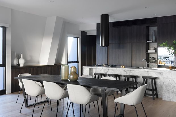Contemporary open-plan kitchen and dining area with a long dark dining table surrounded by upholstered dining chairs. The kitchen features dark timber cabinetry, a marble-look island and splashback, integrated shelving, a black cylindrical rangehood, and light timber flooring. White walls and sculptural angled architectural detailing create a bright, refined interior.