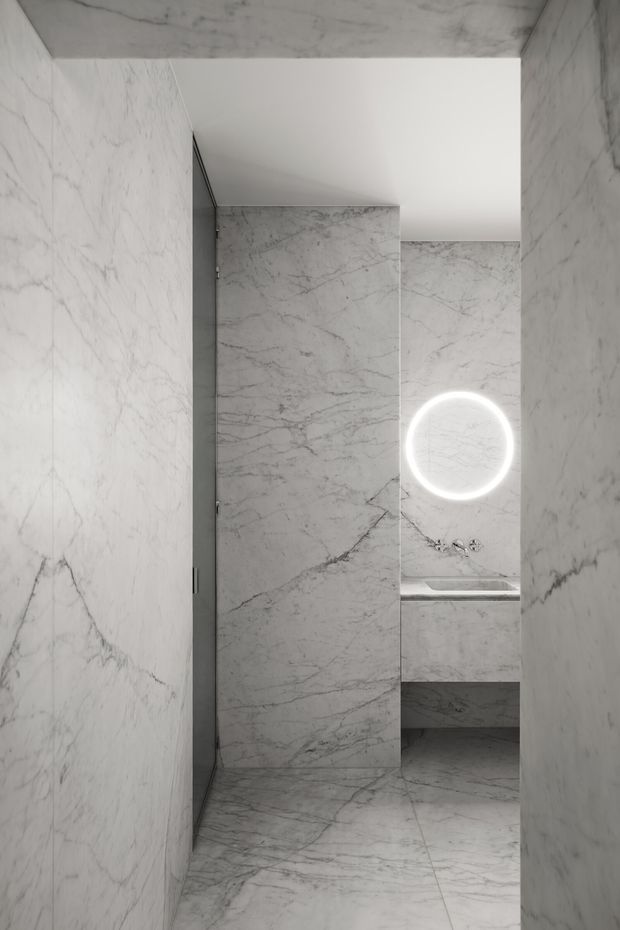 Minimalist bathroom clad almost entirely in white-grey veined marble, viewed through a narrow entry. A freestanding or wall-mounted basin area is visible at the rear with chrome fixtures and a large circular illuminated mirror. The space has a clean, monolithic, highly refined appearance with concealed detailing and a walk-in shower or partitioned wet area.