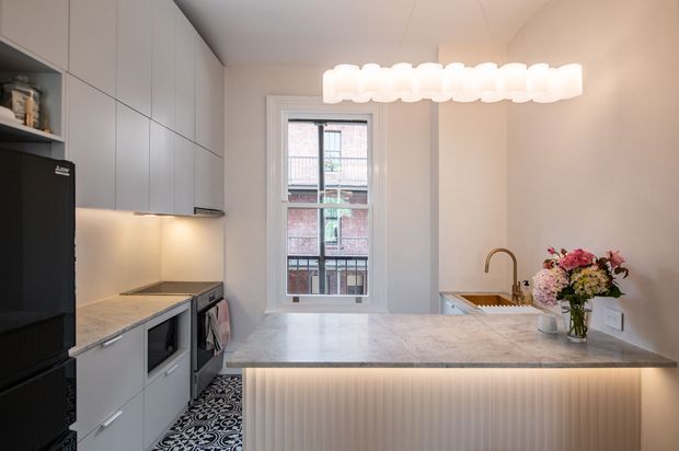 Compact, refined contemporary kitchen with a peninsula island, pale stone benchtop, integrated light-grey cabinetry, black appliances, patterned floor tiles, and a sculptural white pendant light. A window provides natural light, while a fluted illuminated island front and brass-toned tap add detail.