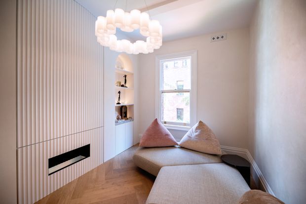 A compact, bright living room with a contemporary built-in fireplace wall featuring vertical light-toned panelling, integrated shelving, and a concealed low fireplace. A pale L-shaped sectional sofa occupies the room, with textured neutral cushions, warm herringbone timber flooring, a tall sash-style window, and a sculptural white ring pendant light.