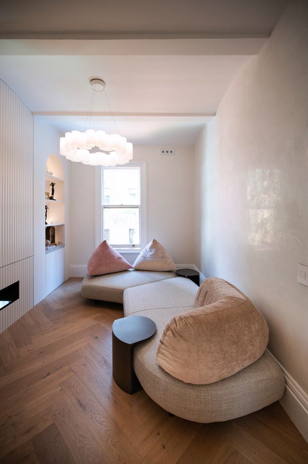 A compact, bright living room with a low modular beige sectional sofa, sculptural pendant light, pale textured walls, warm timber parquet flooring, and built-in white cabinetry with vertical fluted detailing. A narrow window and soft neutral cushions create an airy, refined atmosphere.