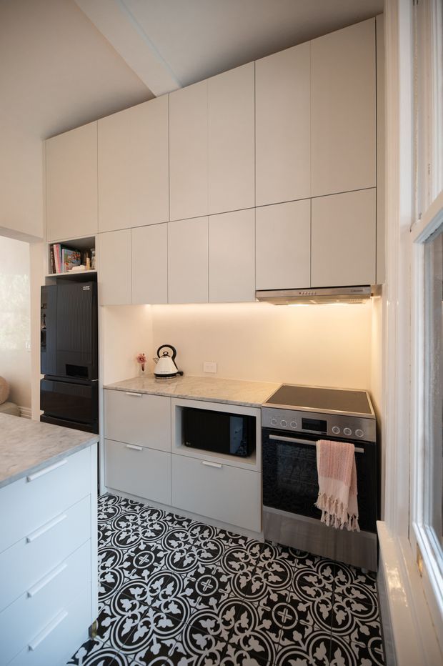 Compact contemporary kitchen with full-height pale cabinetry, integrated under-cabinet lighting, a marble-look stone benchtop, stainless steel oven and cooktop, dark refrigerator, and a bold black-and-white patterned tile floor. A large window provides natural light, while a peninsula with drawers is visible in the foreground.