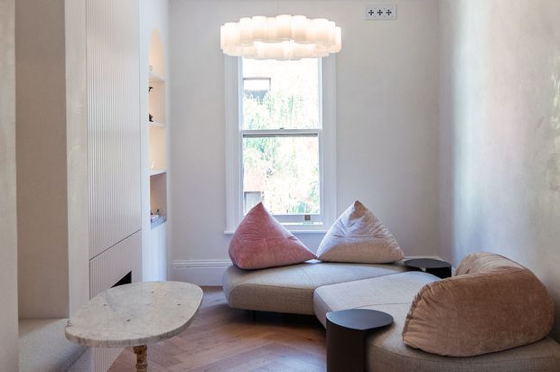 A compact, softly styled living room with a curved sectional sofa, sculptural lounge chairs, small side tables, a pale stone or terrazzo coffee table, timber parquet flooring, built-in wall joinery, and a large central window. A layered white pendant light hangs overhead, creating a warm, refined atmosphere with neutral upholstery and blush-toned cushions.