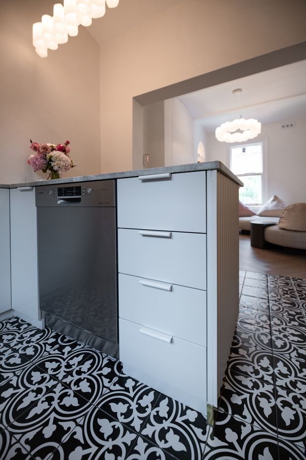 A compact kitchen viewed from the work area, featuring a white island with integrated drawers, a stainless-steel dishwasher, a grey stone-look benchtop, and striking black-and-white patterned floor tiles. A floral arrangement decorates the island, while an adjoining living room with a sofa and sculptural pendant lighting is visible beyond the wide opening.