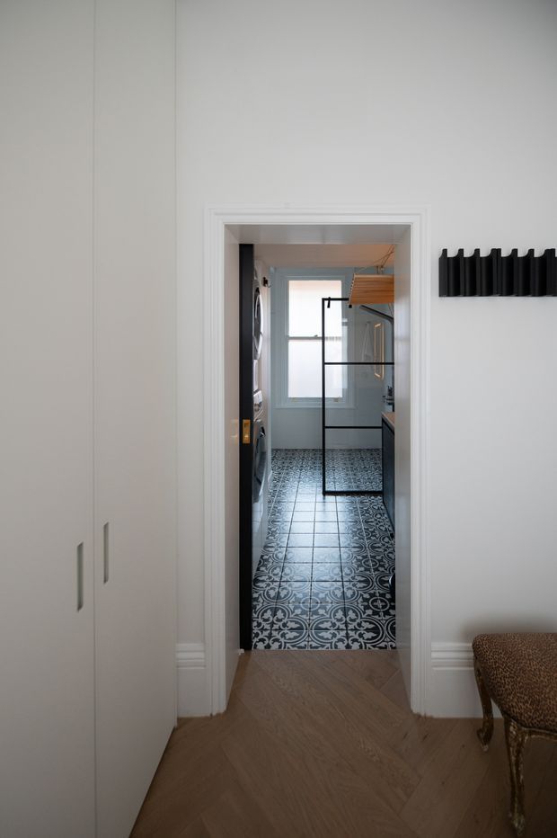 A narrow interior view from a hallway or entry area into a compact laundry room. The foreground has pale built-in cabinetry, white walls with decorative trim, and a warm herringbone timber floor. Beyond the doorway, the laundry features patterned black-and-white floor tiles, dark appliances on the left, pale cabinetry and benchtops on the right, a frosted window, and a black-framed glass partition or door. A sculptural black wall-mounted coat rack and part of a traditional upholstered chair are visible in the foreground.