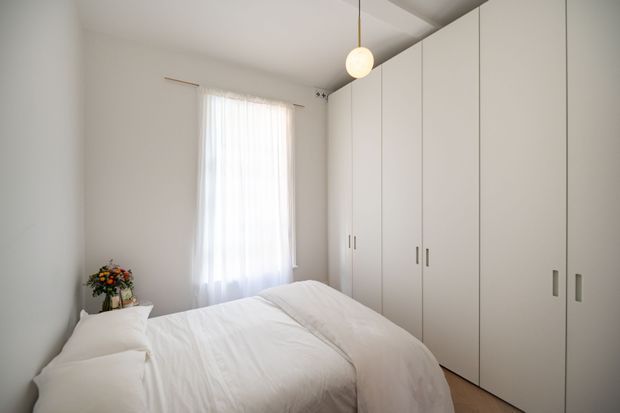 Bright, minimalist bedroom with a neatly made bed, sheer white curtain over a tall window, and extensive floor-to-ceiling white built-in wardrobes with simple vertical handles. A small floral arrangement sits beside the bed, and a spherical pendant light hangs from the ceiling. The room has a calm, uncluttered appearance with light timber flooring.