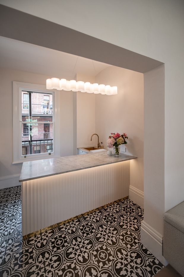A compact, refined kitchen framed by a wide opening, featuring a marble-look peninsula with a vertically fluted white island base, integrated under-counter lighting, a small undermount sink with brass-toned tap, and a decorative white globe pendant. The room has pale walls and trim, a black-and-white patterned encaustic-style tile floor, and a tall window overlooking neighbouring brick buildings.