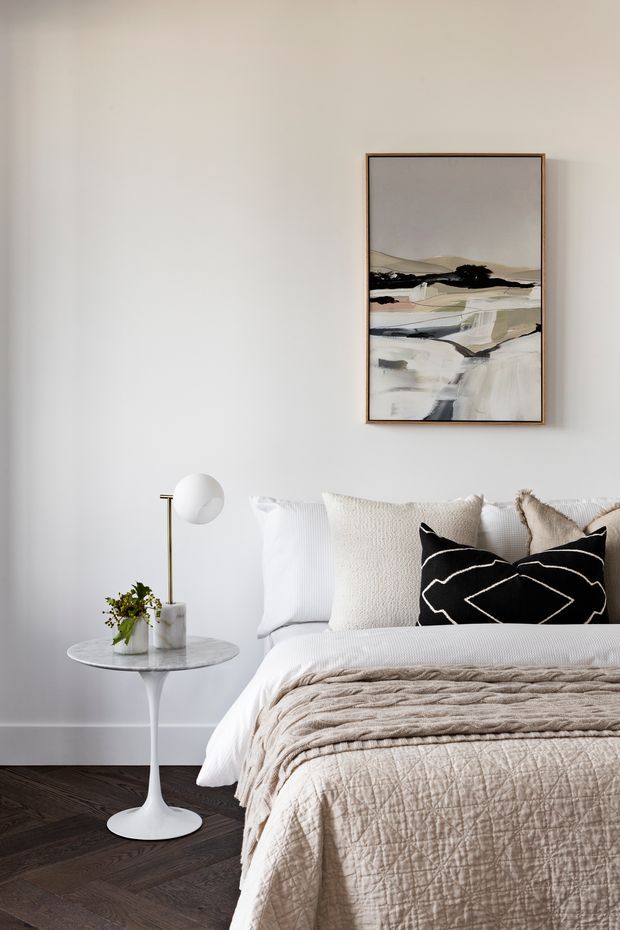 A softly styled bedroom with a neatly made bed, layered cream and beige bedding, textured neutral pillows, and a black accent cushion. The room has pale walls, dark herringbone timber flooring, a slim white side table with a marble-like top, and a simple globe floor lamp. A framed abstract landscape artwork hangs above the bed, creating a calm, refined, contemporary-neutral interior.