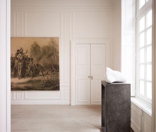 A bright, refined interior gallery or formal hallway with white paneled walls, ornate double doors, pale timber flooring, and tall multi-pane windows. A large sepia artwork hangs on the wall beside a dark pedestal displaying a white sculptural object. The space has a restrained, museum-like atmosphere with classical architectural detailing and abundant natural light.