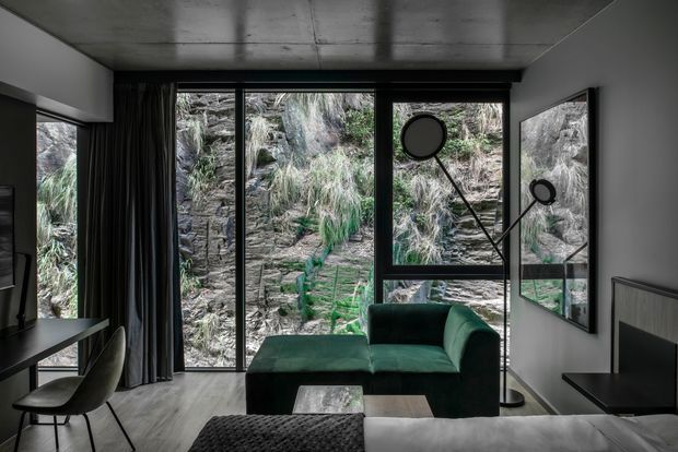 A compact, dark-toned living room with a deep green sectional sofa positioned before full-height black-framed glazing. The glazing overlooks a rugged rocky landscape with grasses and vegetation. A concrete ceiling, dark timber flooring, black-framed windows and doors, a wall-mounted television, sculptural floor lamp, side table, and textured furnishings create a refined contemporary interior with a strong connection to nature.