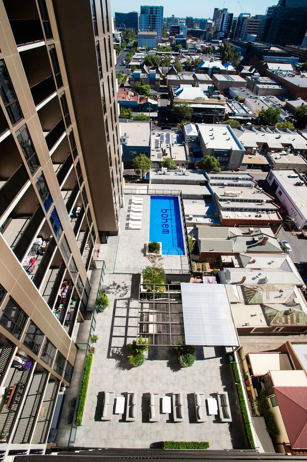 High-angle view from a tall apartment building overlooking a dense urban neighbourhood. The foreground shows a landscaped communal rooftop terrace with pale paved flooring, rows of outdoor loungers, planted areas, a covered pergola-like structure, and a rectangular blue swimming pool marked with lettering. Apartment balconies and facades frame the left side, while low-rise rooftops and taller commercial and residential buildings extend into the city background.