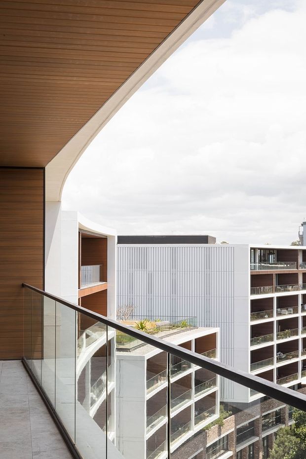 View from a contemporary apartment balcony or terrace toward surrounding multi-storey buildings. The foreground features a glass balustrade with a dark metal handrail, pale tiled flooring, and a dramatic curved architectural canopy lined with warm timber boards. The visible buildings have white and grey façades, vertical metal cladding, extensive glazing, and balconies. Overcast daylight creates a clean, modern urban atmosphere.