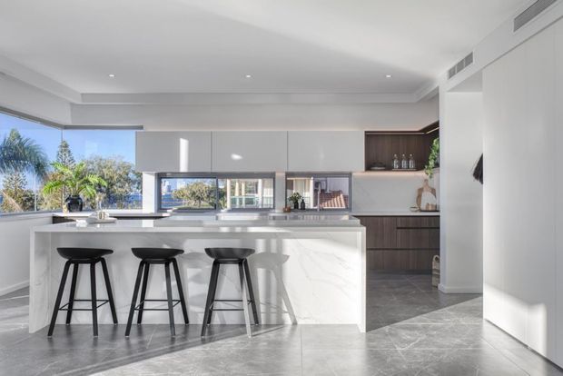 Bright contemporary kitchen interior featuring a large white marble-look island with seating for three, dark bar stools, white flat-panel cabinetry, dark timber-toned joinery, integrated appliances, and large windows overlooking greenery. Grey stone-look flooring and a restrained neutral palette create a clean, refined space.