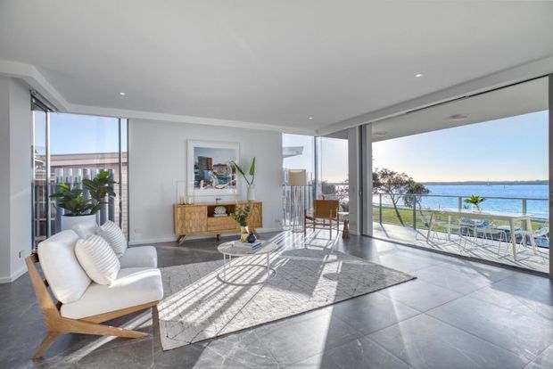 Bright contemporary living room with white walls, expansive floor-to-ceiling glazing, pale tiled flooring, and a large rug. A white upholstered lounge chair, timber sideboard, indoor plants, and minimal furnishings overlook a balcony and blue waterfront beyond.