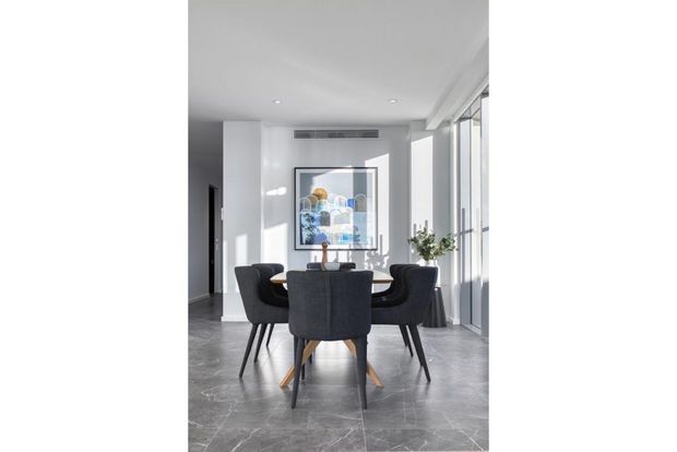 Bright, minimalist dining area with a dark rectangular dining table and upholstered dining chairs, set against white walls and large floor-to-ceiling windows. Grey stone-look flooring, recessed ceiling lights, a framed artwork, and a potted plant complete the contemporary interior.