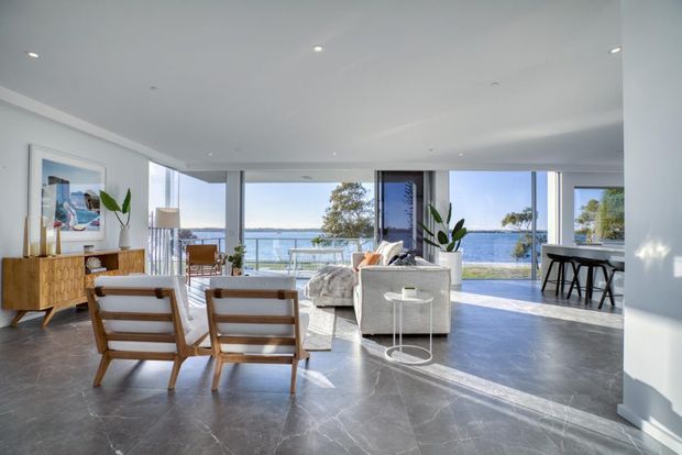 Bright, spacious open-plan living room with contemporary furnishings, polished grey stone flooring, white walls and ceiling, and extensive floor-to-ceiling glazing opening to a waterfront view. The room includes a timber sideboard, lounge seating, armchairs, indoor plants, and a compact bar or kitchen counter along the right side.