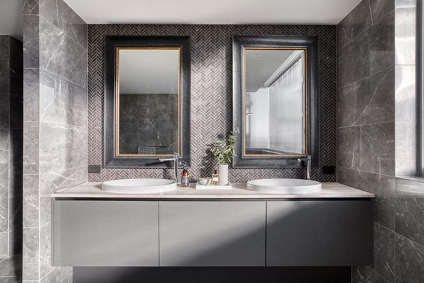 A refined contemporary bathroom featuring a wide floating double vanity with two integrated basins, dark grey stone-look wall tiles, and a central herringbone-pattern feature wall. Two black-framed mirrors with brass-toned inner trim sit above the vanity, complemented by wall-mounted taps and a bright window on the right.