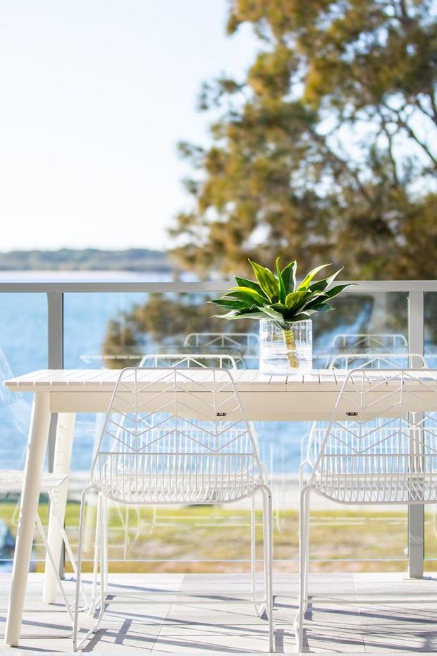 Bright coastal outdoor dining area on a balcony or terrace, furnished with a white rectangular table and matching wire-frame bar chairs. A potted green plant forms a centrepiece, with a glass balustrade overlooking blue water and a tree-lined shoreline.