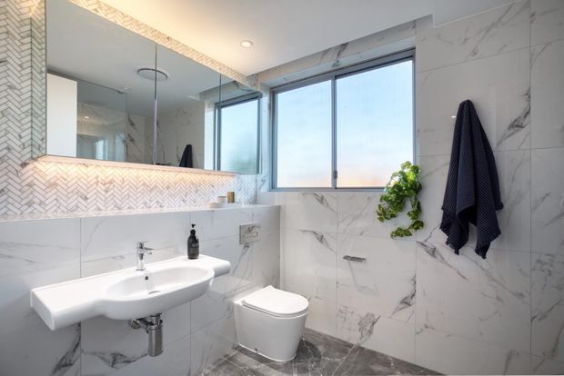 Bright contemporary bathroom featuring large white marble-look wall tiles, a grey marble-look floor, a wall-mounted white basin with chrome tapware, a compact toilet, and a large mirrored vanity wall with a herringbone-tile feature border. A broad window provides natural light, while a small hanging plant and dark towels add contrast.