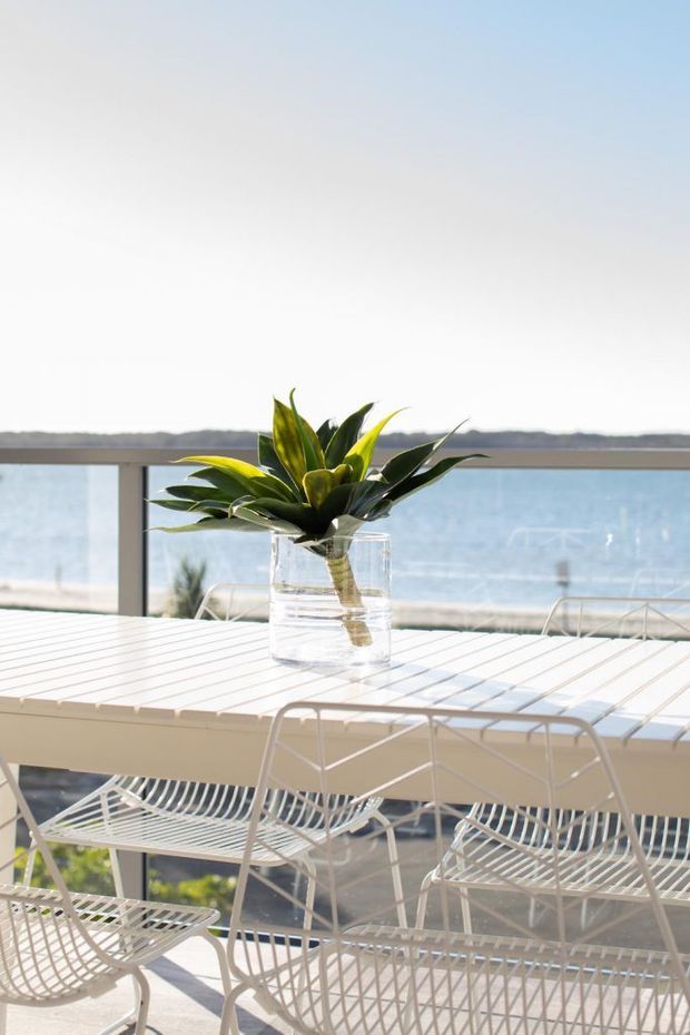 Bright coastal outdoor dining area on a balcony or terrace, furnished with a white slatted dining table and matching wire-frame chairs. A potted tropical plant is the centrepiece, with a glass balustrade and expansive blue water view beyond. The scene has a light, airy resort-like atmosphere.