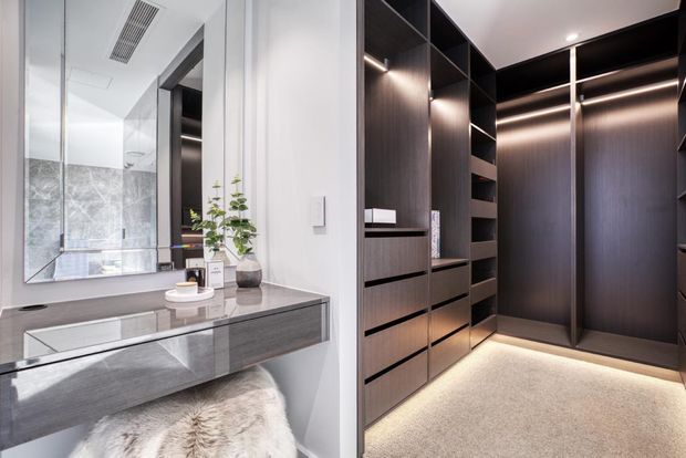 Contemporary dressing and vanity area featuring dark timber built-in wardrobe cabinetry with open shelving, drawers, integrated strip lighting, and a softly illuminated hanging section. A grey stone-look vanity with mirror, basin, and decorative plant sits beside the wardrobe, with a plush light rug adding softness.