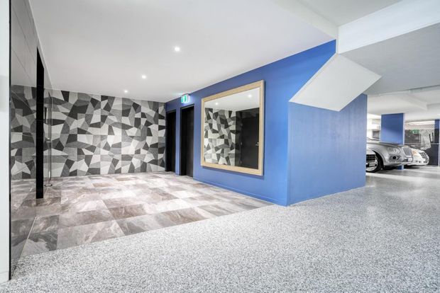 A modern building entry or lobby adjoining a parking garage. The space features vivid blue painted walls, a large framed mirror, a geometric black-and-white patterned feature wall, pale tiled flooring, recessed ceiling lights, and a grey speckled garage floor. Parked vehicles are visible through the opening on the right.