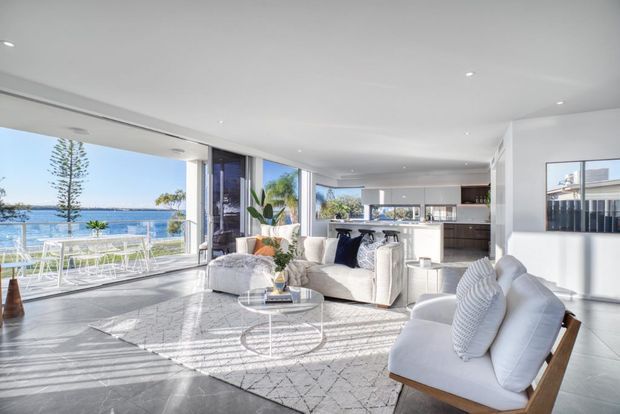 Bright, spacious open-plan living room with floor-to-ceiling glazing overlooking a body of water. The room features white sofas and armchairs, a round glass coffee table, a pale geometric rug, a white kitchen with island, timber accents, and abundant natural light. A balcony or terrace is visible beyond the glazing.