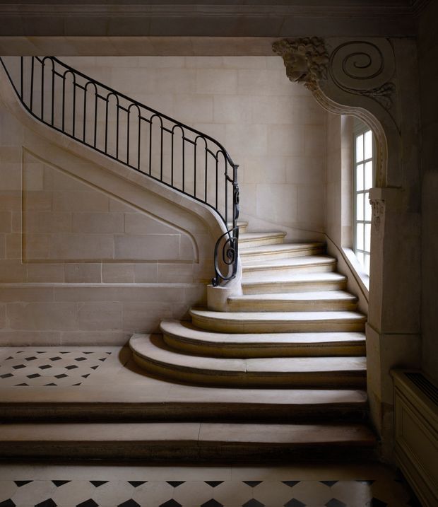 An ornate curved staircase in a grand classical interior, featuring pale stone or plaster walls, broad cream-colored stair treads, an elegant black wrought-iron balustrade, decorative carved scrollwork, panelled detailing, and a tall multi-pane window. The floor has a geometric black-and-white patterned surface, creating a refined heritage atmosphere.