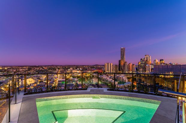 Rooftop or elevated outdoor pool terrace overlooking a brightly illuminated urban skyline at dusk. The foreground features a curved, light-filled swimming pool with surrounding paved decking and transparent glass balustrades. High-rise buildings and city lights form the distant backdrop under a deep blue-purple sky.