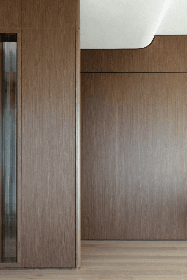 Close-up interior view of refined warm timber wall panelling and full-height built-in joinery, with a narrow fluted glass insert at left, pale timber flooring, and a sculptural white curved ceiling or bulkhead detail. The composition is minimal and highly detailed, focused on materiality and seamless cabinetry.