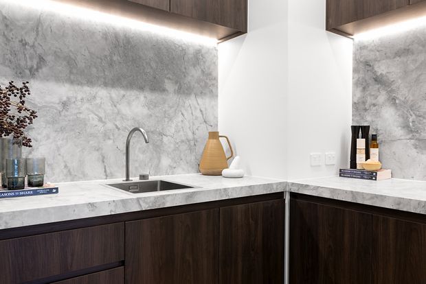 A refined contemporary kitchen or kitchenette with dark wood cabinetry, a pale veined stone benchtop and matching full-height splashback. The compact L-shaped layout includes an undermount sink with a slim stainless-steel tap, warm under-cabinet lighting, white walls, and carefully styled accessories.