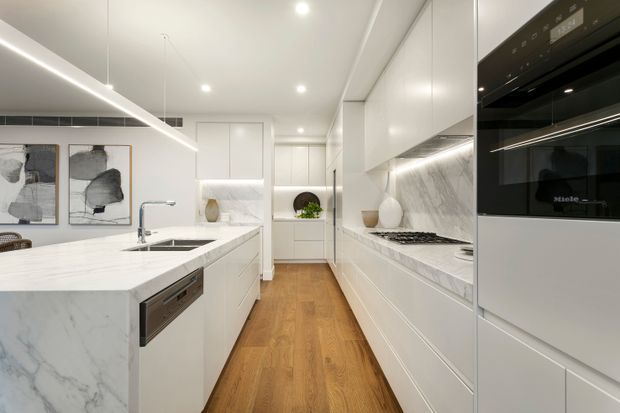 Bright contemporary galley kitchen with a large marble-look island, integrated sink and dishwasher, white handleless cabinetry, marble-look benchtops and splashback, black integrated appliances, warm timber flooring, and linear under-cabinet and ceiling lighting. The space appears compact to medium-sized with a standard-height ceiling and a clean, refined aesthetic.
