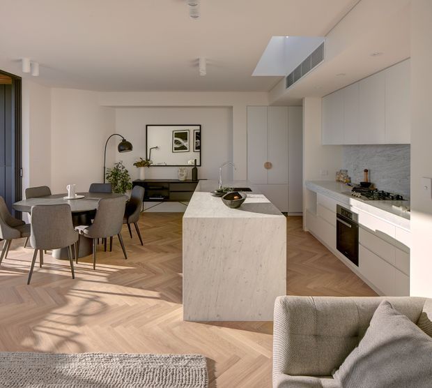 Bright open-plan apartment interior combining a contemporary white kitchen, central stone island, dining area, and living space. The kitchen features flat-panel white cabinetry, a pale veined stone benchtop and splashback, integrated appliances, and a concealed rangehood. Warm herringbone timber flooring runs throughout, with neutral upholstered furniture, soft natural light, and minimalist black and timber accents. A skylight or rooflight above the kitchen contributes to the airy atmosphere.