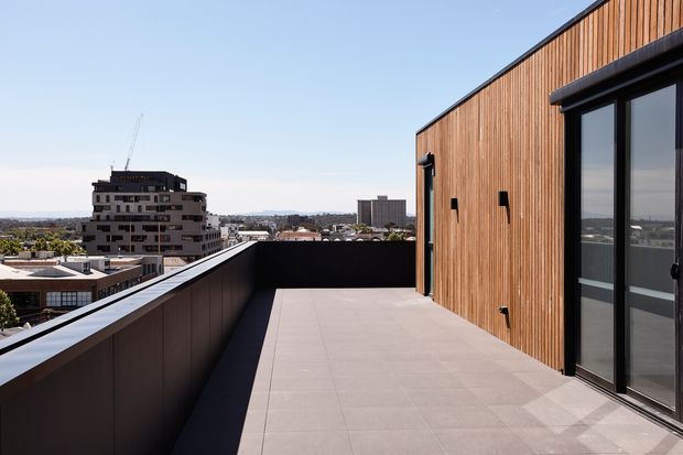 A spacious rooftop terrace or balcony on a modern multi-storey building, with large pale grey tiled paving, dark metal balustrades, and a warm vertical timber-clad exterior wall with black-framed glazed doors. An urban skyline is visible beyond the terrace.