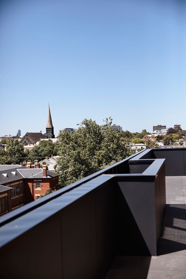 A rooftop terrace or balcony with a dark, angular metal balustrade and grey tiled paving, overlooking a leafy urban neighbourhood. A prominent historic church spire, brick residential buildings, and mid-rise city buildings are visible beyond the terrace under a clear blue sky.
