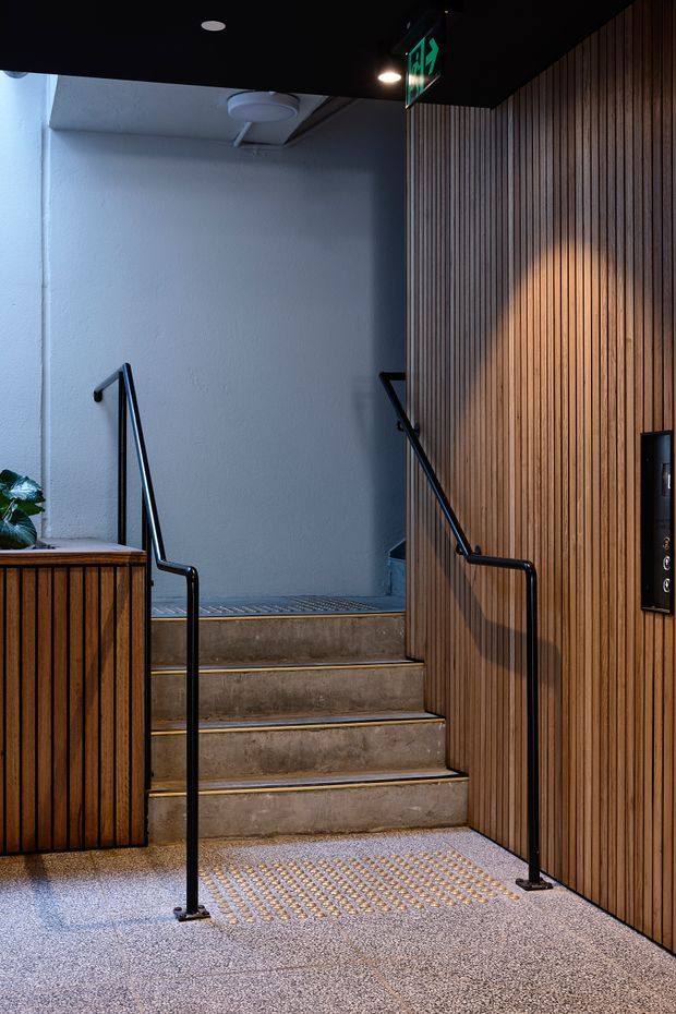 A contemporary commercial or public building stairwell and access area. Concrete steps with yellow edge strips lead upward between black metal handrails. Vertical timber slat wall panelling lines the right side and a matching timber-clad counter or partition sits to the left. The floor has a textured tactile surface, with an illuminated exit sign and understated ceiling lighting overhead. The space is dimly lit but visually refined, combining warm timber with cool grey concrete and white plaster.