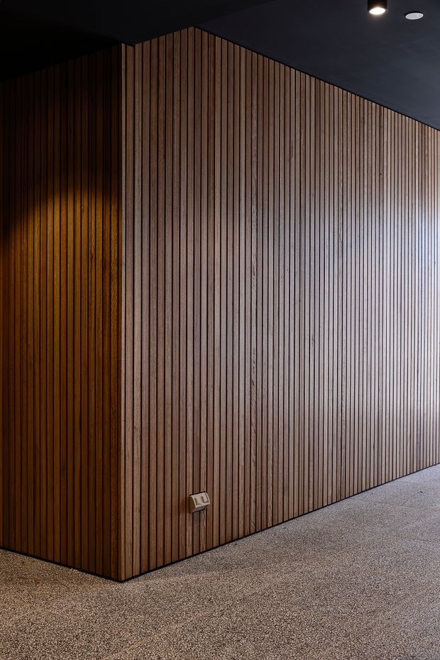 A dark, minimalist interior corner dominated by a full-height vertical timber slat wall wrapping around a projecting volume. Warm concealed lighting grazes the darker side wall, while recessed and surface-mounted ceiling lights illuminate the space. The floor is a speckled grey finish, and a small wall outlet is visible near the base. No furnishings are present.