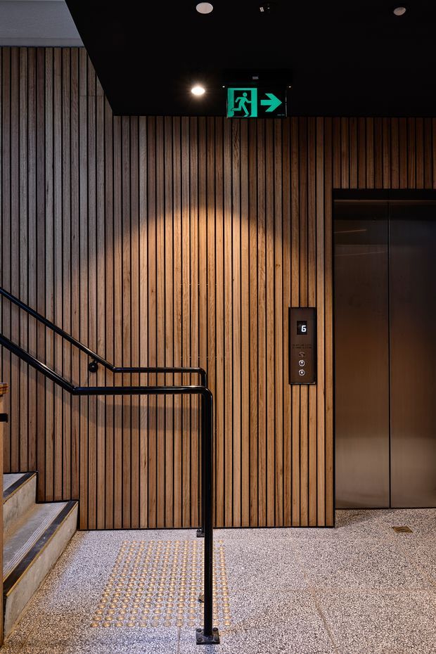 A dimly lit contemporary elevator lobby and stair landing. The walls are clad in vertical warm timber battens, with a black ceiling, recessed downlights, a dark metal handrail, bronze-toned elevator doors and call panel, and a terrazzo-like speckled floor with tactile stair indicators. A green emergency exit sign is mounted above the wall.