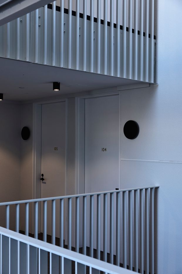 A minimalist multi-level interior corridor or apartment landing with pale grey-white walls, numbered doors, black circular window or feature openings, and repeated light-grey metal balustrades. The view focuses on the clean geometric composition of the walkway and upper railing, with recessed black ceiling lights and subtle wall panelling.