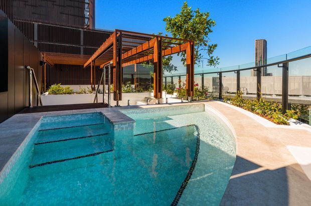A bright outdoor pool area on an urban rooftop or elevated terrace, featuring a turquoise swimming pool with lane markings, a shallow entry or spa section, pale paving, landscaped planter beds, glass safety balustrades, and a timber pergola with orange-brown structural framing. A contemporary building facade and city structures are visible beyond.