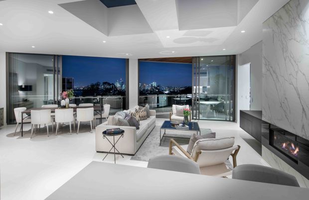 A bright, high-end open-plan living and dining area with expansive floor-to-ceiling glazing overlooking a nighttime city skyline. The room features white furniture, a pale floor, sculptural recessed ceiling forms, a marble-look feature wall with a linear fireplace, and a compact dining setting near the windows. A glazed outdoor terrace or balcony is visible beyond the living area.